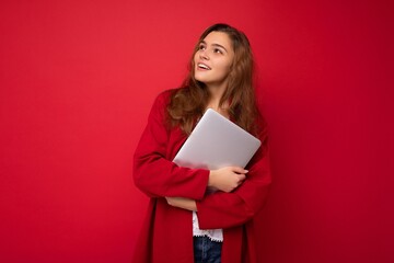 Fototapeta premium shot of thoughtful mysterious Beautiful young dark hair curly woman holding close laptop wearing red cardigan and white blouse looking to the side isolated over red background