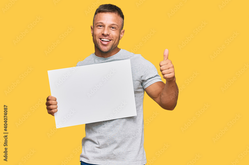 Poster Handsome muscle man holding blank empty banner smiling happy and positive, thumb up doing excellent and approval sign