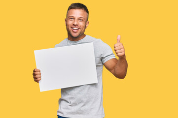 Handsome muscle man holding blank empty banner smiling happy and positive, thumb up doing excellent and approval sign