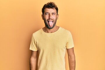 Handsome man with beard wearing casual yellow tshirt over yellow background sticking tongue out happy with funny expression. emotion concept.