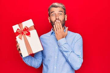 Young handsome blond man with beard holding birthday present over isolated red background covering...