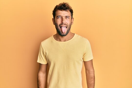 Handsome man with beard wearing casual yellow tshirt over yellow background sticking tongue out happy with funny expression. emotion concept.
