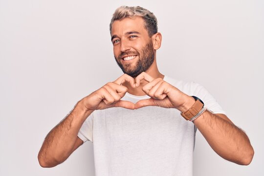 Young handsome blond man wearing casual t-shirt standing over isolated white background smiling in love doing heart symbol shape with hands. Romantic concept.