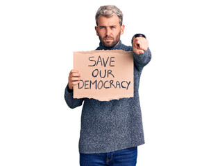 Young blond man holding save our democracy cardboard banner pointing with finger to the camera and to you, confident gesture looking serious
