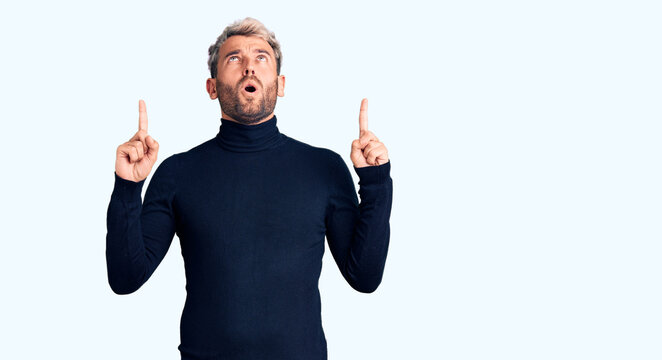 Young handsome blond man wearing casual turtleneck sweater amazed and surprised looking up and pointing with fingers and raised arms.