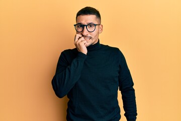 Fototapeta premium Handsome man with tattoos wearing turtleneck sweater and glasses looking stressed and nervous with hands on mouth biting nails. anxiety problem.