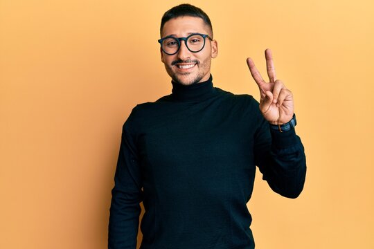 Handsome man with tattoos wearing turtleneck sweater and glasses smiling with happy face winking at the camera doing victory sign with fingers. number two.