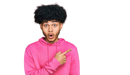 Young african american man with afro hair wearing casual pink sweatshirt surprised pointing with finger to the side, open mouth amazed expression.
