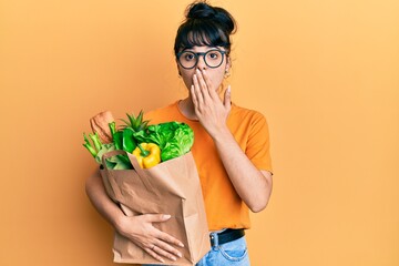 Young hispanic girl holding paper bag with bread and groceries covering mouth with hand, shocked...