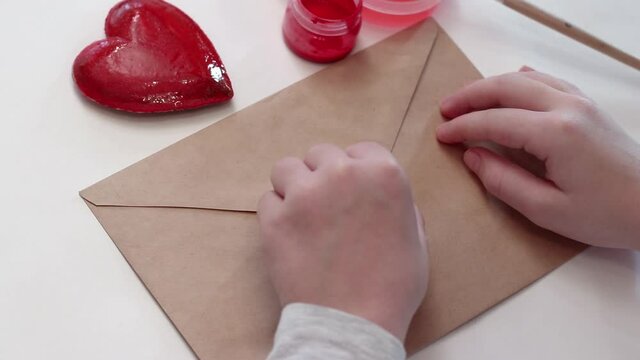 The child paints a decorative heart with red paint and makes an impression on the envelope. Preparing for Valentine's Day.