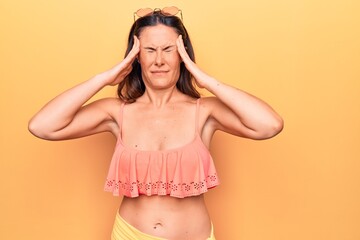 Young beautiful brunette woman wearing bikini standing over isolated yellow background with hand on head, headache because stress. Suffering migraine.