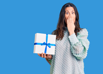 Young beautiful brunette woman holding birthday gift covering mouth with hand, shocked and afraid...