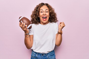 Middle age beautiful sporty woman playing rugby holding football ball over pink background screaming proud, celebrating victory and success very excited with raised arm