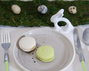 Easter. Serving for the Easter table. A plate of colorful macaroni cookies, cutlery with a green handle on a white tablecloth. Decorative ribbon of green moss and quail eggs.