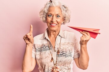 Senior grey-haired woman holding paper airplane smiling with an idea or question pointing finger with happy face, number one