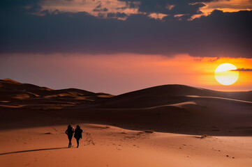 Fototapeta na wymiar (Selective focus) Silhouette of two people walking on the sand dunes of the Merzouga desert during a stunning sunset. Merzouga, Morocco.