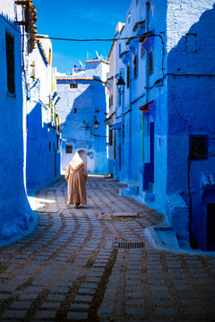 (Selective focus) A man, wearing the Djellaba or Jillaba, is walking throughth the streets of the blue city of Chefchaouen, Morocco. 