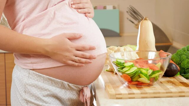Closeup of pregnant woman with big belly standing on kitchen and touching big abdomen. Concept of healthy lifestyle and nutrition during pregnancy