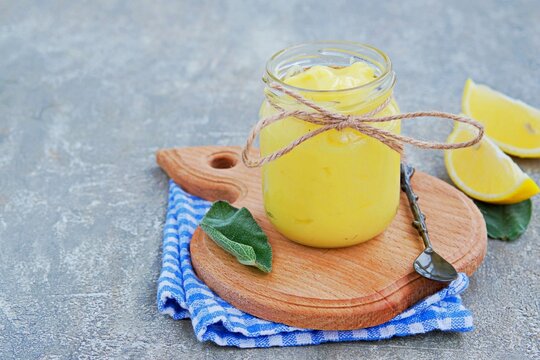 Lemon curd, a sweet fruity custard in a glass jar on a gray concrete background. Cream recipes.