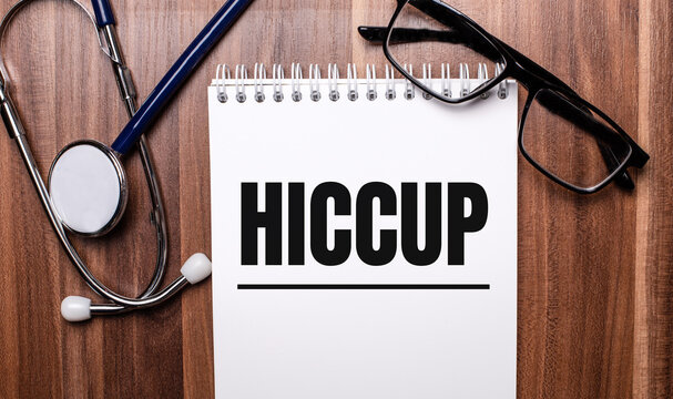 The word HICCUP is written on white paper on a wooden background near a stethoscope and black-framed glasses. Medical concept