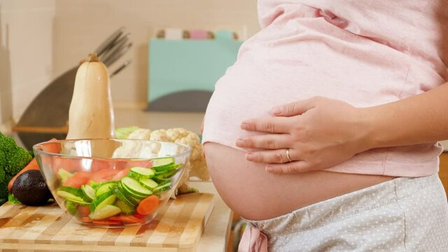 CLoseup of young pregnant woman cooking and eating vegetable salad holding big abdomen and touching it with hands