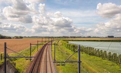 Curved double track train rails with gantries with electric catenaries in a Dutch agricultural landscape. On the right are fruit trees covered with plastic film and wind turbines are in the background