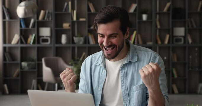 Excited young business man sit at desk read good news on laptop, male winner celebrate online bid game win, career advance, got monetary reward, success and moment of victory candid emotions concept