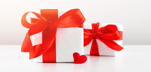 Gift box with red bow ribbon and red paper heart on white table for Valentines day.