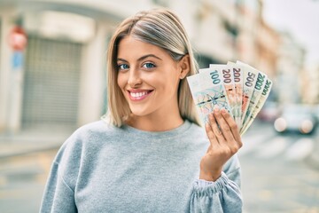 Young blonde girl smiling happy holding czech koruna banknotes at the city.