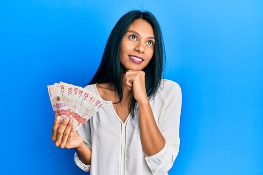 Young african american woman holding 10 colombian pesos banknotes with hand on chin thinking about question, pensive expression. smiling and thoughtful face. doubt concept.