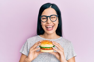 Beautiful young woman eating tasty hamburger winking looking at the camera with sexy expression, cheerful and happy face.