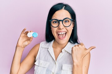 Beautiful young woman holding glasses and contact lenses pointing thumb up to the side smiling...