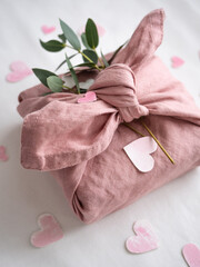 Gift wrapped in pink linen decorated with eucalyptus on a background of paper pink hearts on a white background in the morning light