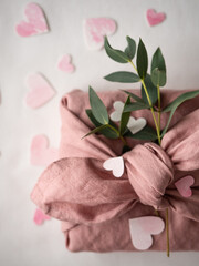 Gift wrapped in pink linen decorated with eucalyptus on a background of paper pink hearts on a white background in the morning light