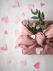 Gift wrapped in pink linen decorated with eucalyptus on a background of paper pink hearts on a white background in the morning light