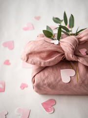 Gift wrapped in pink linen decorated with eucalyptus on a background of paper pink hearts on a white background in the morning light