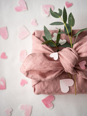 Obraz na płótnie Canvas Gift wrapped in pink linen decorated with eucalyptus on a background of paper pink hearts on a white background in the morning light