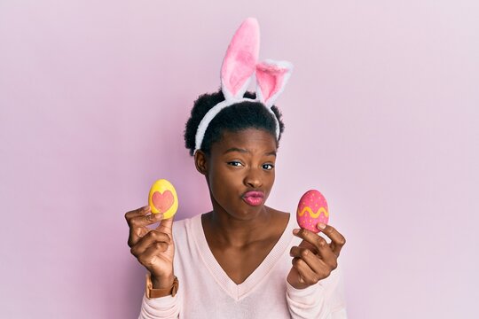 Young african american girl wearing cute easter bunny ears holding painted eggs looking at the camera blowing a kiss being lovely and sexy. love expression.