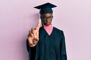 Young african american girl wearing graduation cap and ceremony robe pointing with finger up and angry expression, showing no gesture