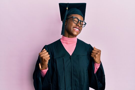 Young african american girl wearing graduation cap and ceremony robe very happy and excited doing winner gesture with arms raised, smiling and screaming for success. celebration concept.