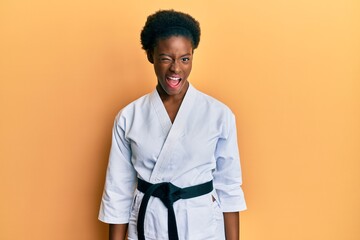 Young african american girl wearing karate kimono and black belt winking looking at the camera with...