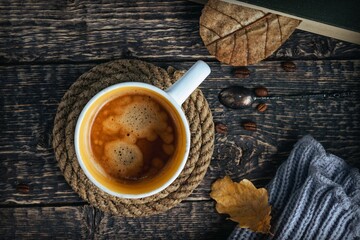 A cup of coffee on a rope coaster, a book, a gray scarf, oak leaves and coffee beans on an old brown wood table.