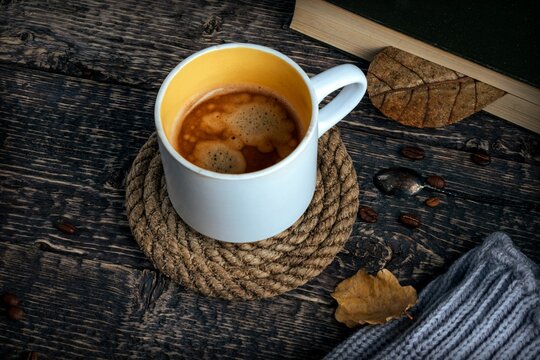 A cup of coffee on a rope coaster, a book, a gray scarf, oak leaves and coffee beans on an old brown wood table.