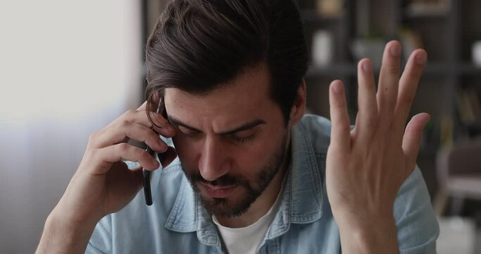 Young man sit indoor talking on smartphone hearing bad news, office employee holding cellphone listen client complaints during phone conversation looking disappointed feel stressed, close up face view