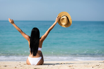 Happy woman in white bikini raises her hands up sitting on beach, sunny day