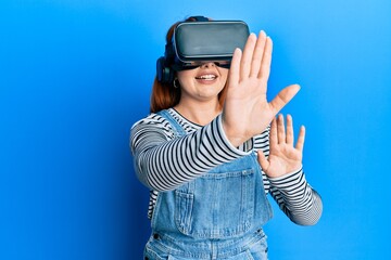 Young redhead woman using 3d virtual glasses standing over isolated blue background.