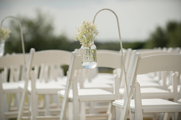 Fancy white folding chairs lined  up at outdoor wedding ceremony with babyie's breathe flowers in mason jars hanging