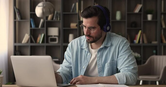 Focused man sit at desk wearing glasses and wireless headphones listens audio course using laptop writing notes studying online. Educational webinar lesson, e-learning, self-education remotely concept