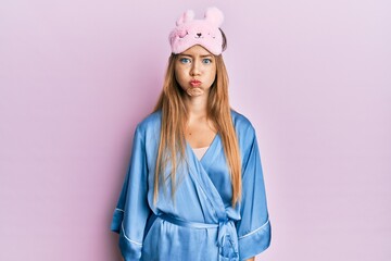 Beautiful young blonde woman wearing sleep mask and pajama puffing cheeks with funny face. mouth inflated with air, crazy expression.