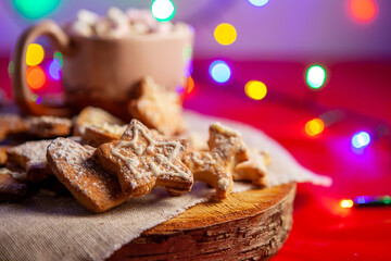 Christmas gingerbread cookies on a wooden stand and a cup of hot chocolate with marshmallows in the back, on a red background with a beautiful bokeh of glowing garland, selective focus. 